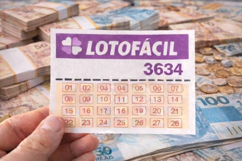 Bilhete da Lotofácil 3634 segurado por mão, com pilhas de dinheiro e moedas em real ao fundo, simbolizando prêmio de loteria.