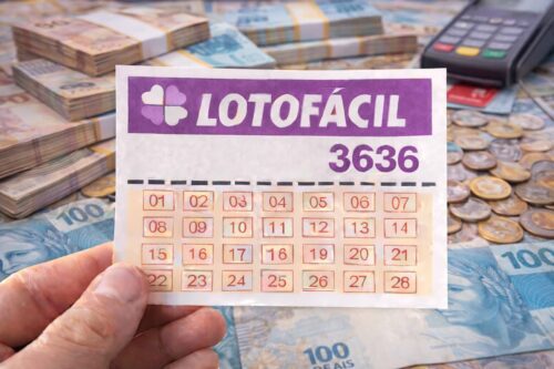 Cartela da Lotofácil 3636 sendo segurada sobre notas e moedas de real, com pilhas de dinheiro e máquina de cartão ao fundo.