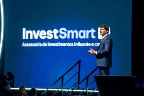 Samyr Castro apresenta palestra no palco do Smart Summit, com telão da InvestSmart ao fundo e plateia assistindo.