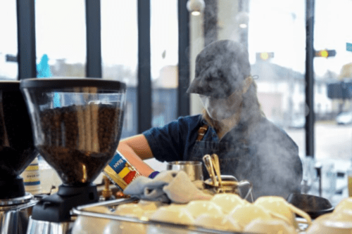 Barista trabalhando em uma cafeteria moderna, vestindo avental, boné e máscara de proteção. Há vapor subindo em frente ao profissional, com moedores de café repletos de grãos em primeiro plano e xícaras organizadas sobre a máquina de espresso. Ao fundo, grandes janelas de vidro revelam a iluminação externa.