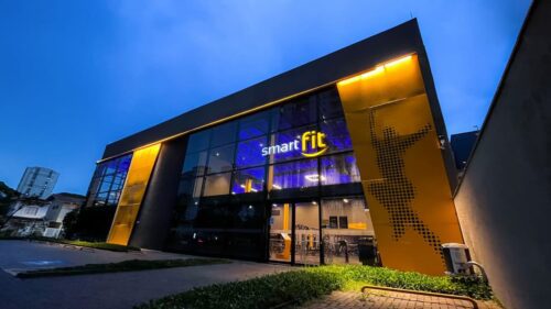 Fachada da academia Smart Fit iluminada à noite, com estrutura moderna e entrada de vidro em destaque.