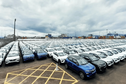 Uma vasta esplanada asfáltica ao ar livre, possivelmente em um porto ou pátio logístico, repleta de centenas de carros novos alinhados em fileiras organizadas. Os veículos são predominantemente modelos SUV compactos da marca Renault, nas cores branco, azul e preto. Ao fundo, sob um céu nublado, veem-se estruturas industriais e guindastes portuários.