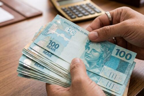 Close-up das mãos de uma pessoa segurando e contando um maço de notas de 100 reais (cedulário brasileiro de cor azul).