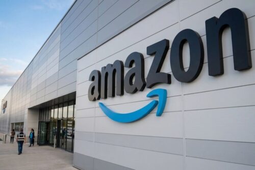 Fachada de um grande centro de distribuição da Amazon com o logotipo da empresa em destaque na parede cinza metálica sob um céu claro.