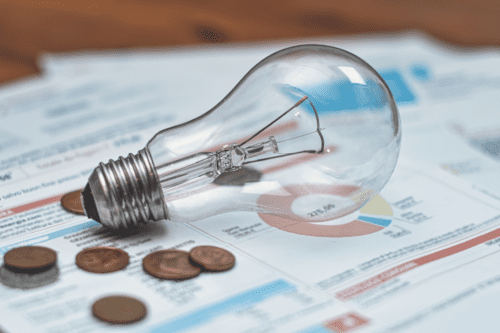 Imagem de uma lâmpada de vidro transparente com base metálica, colocada sobre documentos financeiros com gráficos e alguns moedas ao redor, simbolizando economia e energia.
