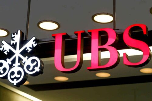 Close-up do letreiro luminoso do banco UBS. As letras "UBS" estão em vermelho tridimensional, acompanhadas pelo logotipo clássico de três chaves cruzadas à esquerda, iluminadas contra um teto interno com luzes embutidas.