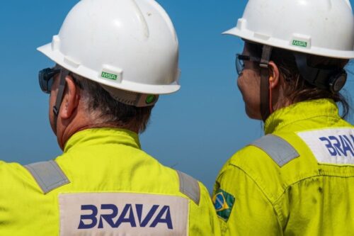 Dois trabalhadores, um homem e uma mulher, vistos de costas e da cintura para cima. Eles vestem uniformes de alta visibilidade na cor amarela vibrante com a logomarca "BRAVA" estampada nas costas e capacetes de proteção brancos.
