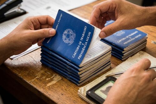 Close-up das mãos de duas pessoas sobre uma mesa de madeira durante um procedimento administrativo. Uma pilha de carteiras de trabalho azuis (CTPS - Brasil) está no centro; uma das pessoas folheia um dos documentos.