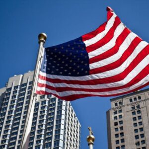 Bandeira dos Estados Unidos da América hasteada e tremulando em destaque no primeiro plano, sob um céu azul límpido. A