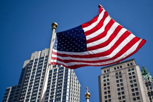 Bandeira dos Estados Unidos da América hasteada e tremulando em destaque no primeiro plano, sob um céu azul límpido. A