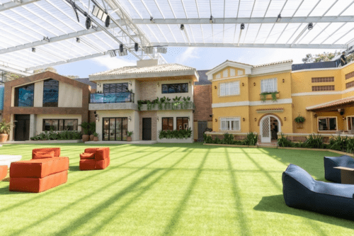 Vista panorâmica da área externa da casa do Patrão. O espaço conta com um amplo gramado artificial verde vibrante, sob uma cobertura retrátil de estrutura metálica e painéis translúcidos que filtram a luz solar, criando sombras lineares no chão.