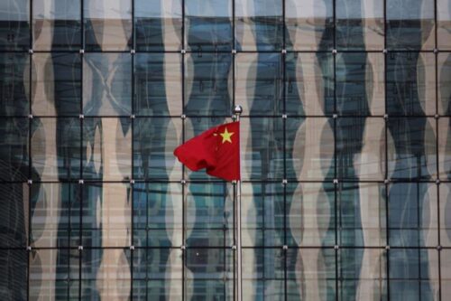 Bandeira da China vermelha com cinco estrelas douradas tremulando em um mastro prateado.