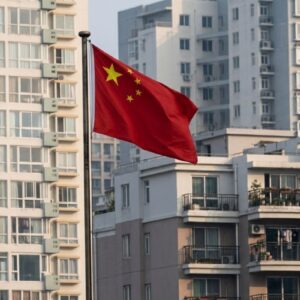 A bandeira nacional da China, vermelha com cinco estrelas douradas, tremula em um mastro no centro da imagem. Ao fundo, há uma paisagem urbana densa composta por prédios residenciais altos e de arquitetura moderna, em tons de bege e cinza, com muitas janelas e varandas visíveis. A iluminação sugere ser a luz do dia, com um céu claro e suave.