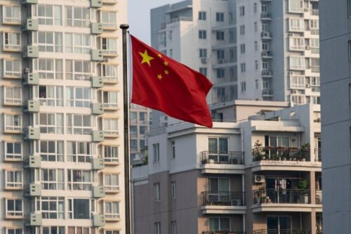 A bandeira nacional da China, vermelha com cinco estrelas douradas, tremula em um mastro no centro da imagem. Ao fundo, há uma paisagem urbana densa composta por prédios residenciais altos e de arquitetura moderna, em tons de bege e cinza, com muitas janelas e varandas visíveis. A iluminação sugere ser a luz do dia, com um céu claro e suave.