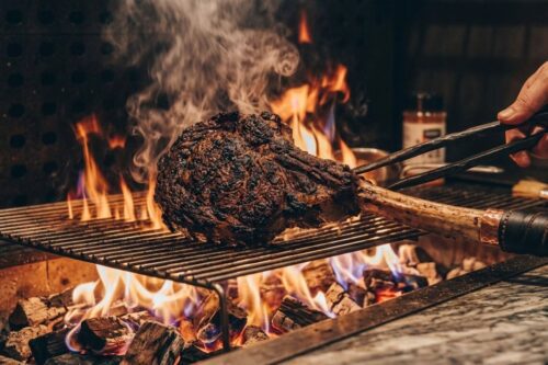 Close-up de um bife de costela (tomahawk) sendo grelhado em uma churrasqueira com fogo alto e fumaça.