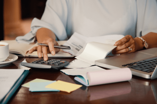 Pessoa trabalhando com documentos, calculadora e computador em uma mesa de escritório, ilustrando tarefas administrativas ou financeiras.