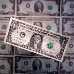 Uma nota de 1 dólar americano (USD) em destaque no centro, levemente inclinada, sobre um fundo composto por várias outras notas de dólar desfocadas, criando um padrão repetitivo da moeda dos Estados Unidos.