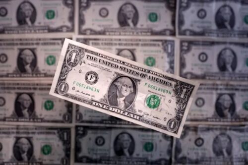 Uma nota de 1 dólar americano (USD) em destaque no centro, levemente inclinada, sobre um fundo composto por várias outras notas de dólar desfocadas, criando um padrão repetitivo da moeda dos Estados Unidos.