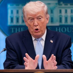 Uma fotografia de médio alcance de Donald Trump no pódio do Salão de Conferências de Imprensa da Casa Branca, falando com as mãos erguidas e palmas para dentro.