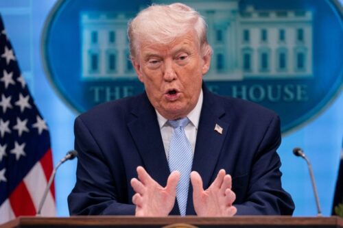 Uma fotografia de médio alcance de Donald Trump no pódio do Salão de Conferências de Imprensa da Casa Branca, falando com as mãos erguidas e palmas para dentro.
