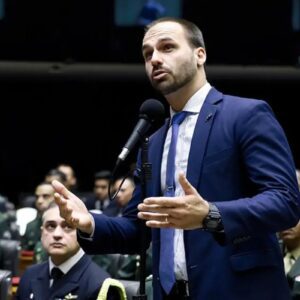 Eduardo Bolsonaro, homem de pele clara, barba curta e terno azul, fala ao microfone em um ambiente legislativo.