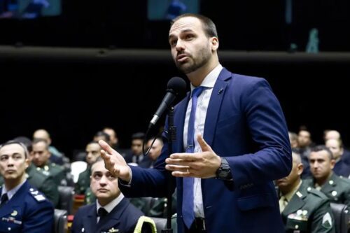 Eduardo Bolsonaro, homem de pele clara, barba curta e terno azul, fala ao microfone em um ambiente legislativo.
