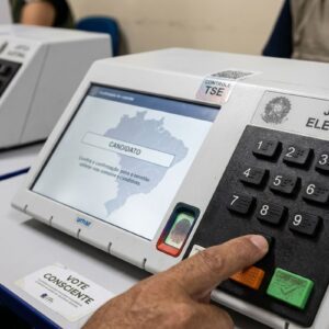 Close-up de uma mão pressionando o teclado numérico de uma urna eletrônica da Justiça Eleitoral brasileira,