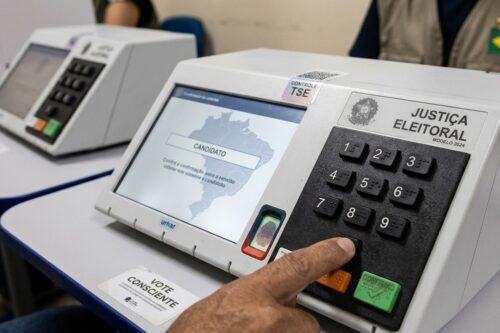 Close-up de uma mão pressionando o teclado numérico de uma urna eletrônica da Justiça Eleitoral brasileira,