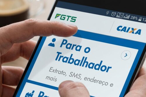 Close de uma mão segurando um smartphone que exibe o aplicativo do FGTS da Caixa Econômica Federal, com o texto "Para o Trabalhador" em destaque na tela.