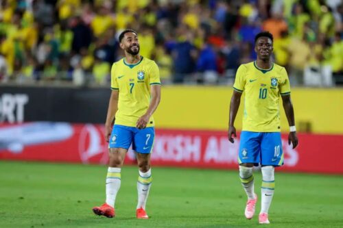 Dois jogadores da Seleção Brasileira de futebol, vestindo a tradicional camisa amarela e calções azuis, caminham em um gramado durante uma partida.