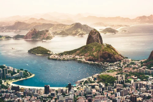 Vista aérea panorâmica da cidade do Rio de Janeiro em um dia ensolarado. No centro, destaca-se o Pão de Açúcar e o Morro da Urca, cercados pelas águas azuis da Baía de Guanabara, onde flutuam dezenas de pequenos barcos brancos. Em primeiro plano, vê-se a densa mancha urbana do bairro de Botafogo com seus prédios e a curva da Praia de Botafogo. Ao fundo, diversas camadas de montanhas se estendem sob um céu claro e levemente amarelado pelo sol, criando um efeito de profundidade.