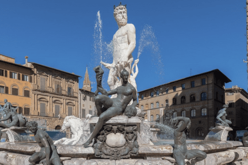 Fotografia em ângulo médio da Fonte de Netuno na Piazza della Signoria, em Florença. No centro, uma imponente estátua de mármore branco de Netuno ergue-se sobre uma carruagem em forma de concha puxada por cavalos marinhos. Ao redor da base da fonte, há várias esculturas de bronze representando divindades marinhas e figuras mitológicas em poses dinâmicas. O fundo mostra edifícios históricos de arquitetura renascentista sob um céu azul limpo.