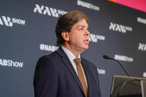 Fotografia de meio corpo do Ministro das Comunicação, Frederico Siqueira Filho, de perfil, falando em um púlpito com microfone. O fundo exibe a marca da feira NAB Show.