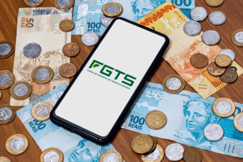 Imagem de dinheiro brasileiro com cédulas e moedas ao redor de um smartphone com o logo do FGTS - Fundo de Garantia do Tempo de Serviço.