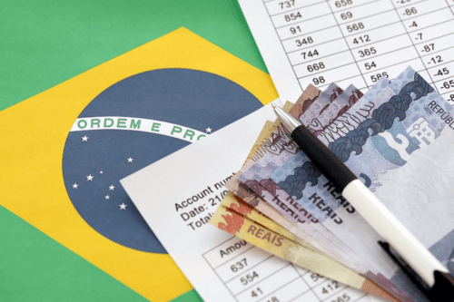 Imagem que demonstra a relação entre o Brasil e o dinheiro, com notas de reais, uma planilha financeira e uma bandeira brasileira ao fundo.