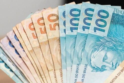 Close em um leque de cédulas de Real brasileiro. À direita, destacam-se notas azuis de 100 reais, mostrando parte da Efígie da República e o texto "República Federativa do Brasil". Ao centro e à esquerda, aparecem notas de 50 reais em tons de bege e laranja, seguidas por notas de valores menores em tons de roxo e azul claro. O fundo é neutro e levemente desfocado, mantendo o foco total no dinheiro.