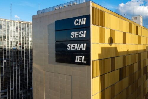 Vista externa de um edifício moderno em Brasília com as siglas CNI, SESI, SENAI e IEL estampadas em uma placa preta sobre a fachada amarela.