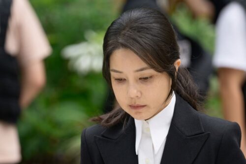 Retrato da ex-primeira-dama Kim Keon Hee, com semblante sério e olhar baixo, vestindo um blazer preto sobre uma camisa branca de gola alta.