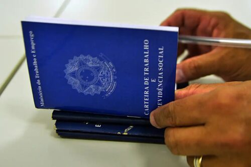 Detalhe das mãos de uma pessoa segurando e manuseando uma Carteira de Trabalho e Previdência Social (CTPS) do Brasil, de cor azul marinho.
