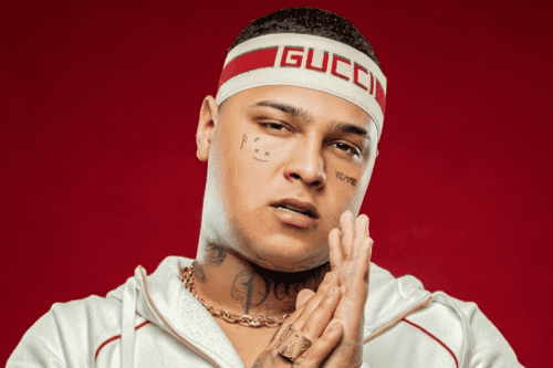 Retrato em close do cantor de funk MC Ryan SP contra um fundo vermelho sólido. Ele usa uma faixa de cabeça branca da marca Gucci com uma listra vermelha central. O artista possui tatuagens no rosto (incluindo um pequeno rosto estilizado abaixo do olho direito) e no pescoço. Ele veste um casaco esportivo branco com capuz e uma corrente de ouro grossa. Ryan está com as mãos juntas à frente do rosto em um gesto de agradecimento ou oração, exibindo um anel de ouro grande no dedo anelar. Ele olha diretamente para a câmera com uma expressão séria.