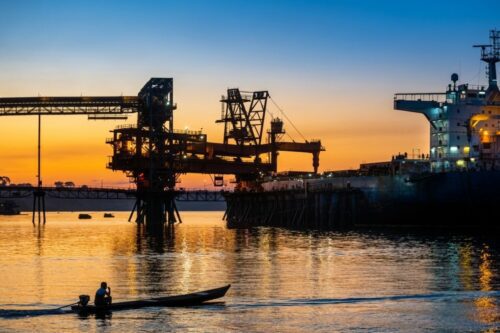Silhueta de um pescador em um pequeno barco motorizado navegando em um rio ao pôr do sol, com um grande navio cargueiro e estruturas portuárias ao fundo.