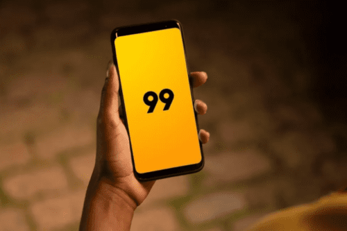 Foto em close das mãos de uma pessoa negra segurando um smartphone. A tela do aparelho está ligada e exibe o logotipo da empresa "99" (o número 99 em preto) sobre um fundo amarelo vibrante. O ambiente ao fundo está levemente desfocado, sugerindo um cenário externo ao anoitecer.