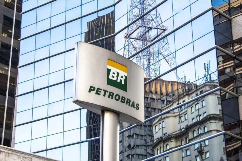 Placa metálica com o logotipo da Petrobras em primeiro plano. Ao fundo, a fachada de um edifício moderno com vidros espelhados azuis que refletem a estrutura de uma torre metálica e parte de uma construção vizinha.