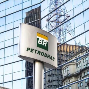 Placa metálica com o logotipo da Petrobras em primeiro plano. Ao fundo, a fachada de um edifício moderno com vidros espelhados azuis que refletem a estrutura de uma torre metálica e parte de uma construção vizinha.