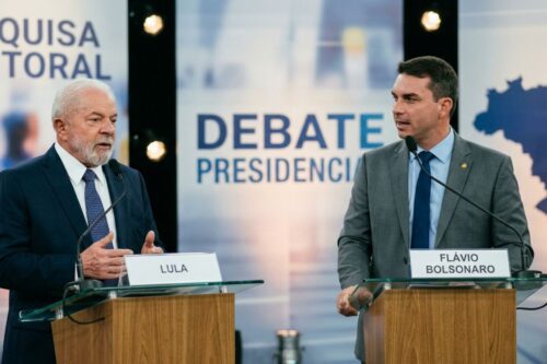 Fotografia de um debate político em estúdio. À esquerda, Luiz Inácio Lula da Silva, vestindo terno azul escuro e gravata estampada, gesticula enquanto fala. À direita, Flávio Bolsonaro, vestindo terno cinza e gravata azul, olha em direção a Lula.