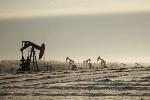 Fotografia de silhuetas de várias unidades de bombeio de petróleo (cavalos-mecânicos) espalhadas por um campo plano sob uma névoa clara e céu pálido.