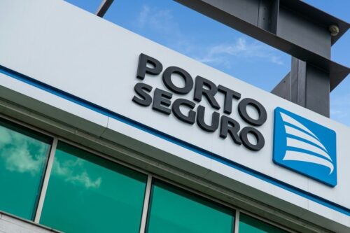 Fachada em ângulo de um prédio moderno exibindo o logotipo da Porto Seguro. O letreiro possui letras pretas em relevo ao lado de um ícone azul quadrado com listras brancas curvadas.