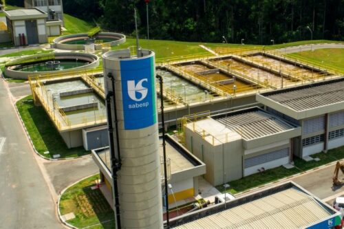 Vista aérea de uma estação de tratamento de água da Sabesp. Em primeiro plano, uma torre cilíndrica cinza com o logotipo azul e branco da empresa.