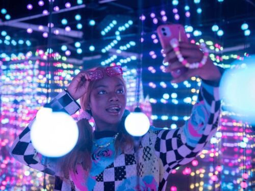 Uma mulher jovem de pele negra sorri alegremente enquanto tira uma selfie em um ambiente imersivo repleto de luzes coloridas. Ela usa óculos de sol cor-de-rosa na testa, um suéter xadrez em tons de preto, branco e azul, e prende o cabelo loiro em dois rabos de cavalo baixos. O fundo é composto por dezenas de pequenas esferas brilhantes em tons de azul, rosa e violeta, criando um efeito visual cintilante e futurista. Ela segura o celular com uma das mãos, que possui uma pulseira de contas coloridas.