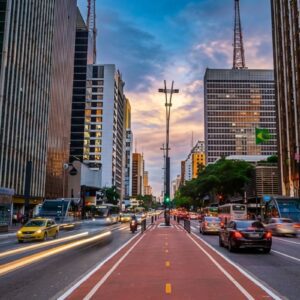 Vista da Ciclovia da Avenida Paulista com rastro de luzes de carros e edifícios ao fundo durante o pôr do sol.